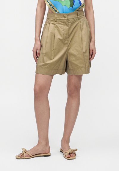 Femme portant un short plissé marron clair, un haut coloré et des sandales beiges à brides nouées, debout devant un fond blanc uni.