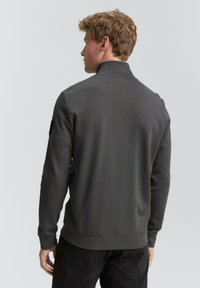 Tmavošedá mikina na zips, ktorá má vysoký golier a pružné manžety, vyrobená z jemného materiálu s jednoduchým dizajnom a logo nášivkou na zadnej strane.