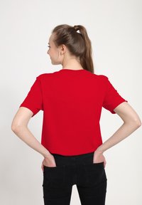 Woman with long brown hair in a ponytail wearing a plain red t-shirt and black jeans with hands in back pockets, facing away on a white background.