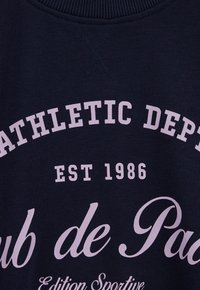 Tengerészkék pulóver pamut anyagból, nagy világos rózsaszín felirattal: "ATHLETIC DEPT EST 1986" és "Club de Paix." Klasszikus környakú kialakítás.