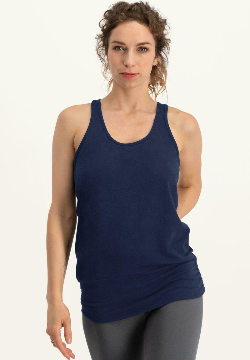 Tummansininen hihaton toppi, jossa on pyöreä kaula-aukko ja kerätty helma, valmistettu pehmeästä kankaasta, yhdistetty harmaisiin tiukkoihin legginsseihin.