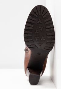 Semelle de botte en cuir marron avec des crampons profonds et la taille 37 inscrite, présentée en position verticale sur un fond blanc.