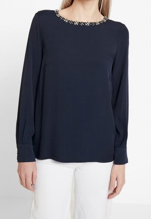 Kobieta ubrana w granatową bluzkę z długim rękawem ozdobioną koralikami wzdłuż dekoltu oraz białe spodnie, stojąca na jednolitym tle.