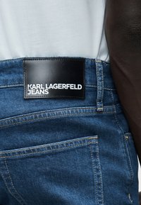 Πίσω όψη ατόμου που φοράει μπλε τζιν τζιν με μαύρη δερμάτινη ετικέτα «Karl Lagerfeld Jeans» στη μέση, και λευκό πουκάμισο που είναι μαζεμένο μέσα.