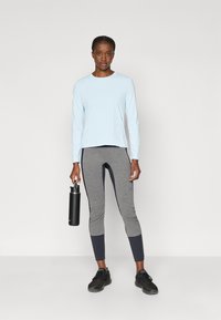 Hele sinine pikakäistega topp on kombineeritud hallide ja meresiniste retuusidega, käes must veepudel ning jalas mustad sportlikud jalanõud.