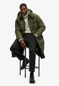 Olivengrøn polstret jakke med hætte, beige sweater, sorte bukser og snørebåndsbukser i sort. Jakken har lynlås og snoretræk.