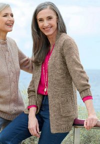 Zwei lächelnde Frauen im Freien in der Nähe von Wasser, die gestrickte Pullover tragen, eine in Beige mit Jeans und einem pinken Hemd darunter, die andere in einem hellbraunen Pullover.