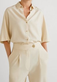 Beige zijden shirt met een kraag en knoopsluiting, gecombineerd met bijpassende geplooide broeken met een gouden knoopdetail op de tailleband.