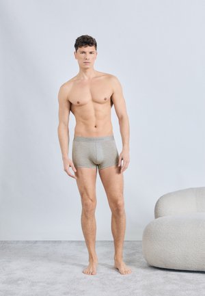Jonge man staat op blote voeten en draagt grijze Calvin Klein boxershorts, tegen een simpele lichtgrijze achtergrond, naast een witte ronde stoel.