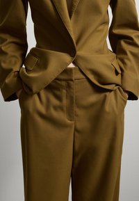 Blazer marrone su misura con chiusura a un bottone abbinato a pantaloni coordinati. Tessuto liscio con forma strutturata e tasche laterali.