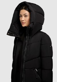 Juoda pūkinė striukė su dideliu gobtuvu, su siuvinėta dizaine, dviem užtrauktukais ir reguliuojamais raišteliais. Yra kišenės ir lygi tekstūra.