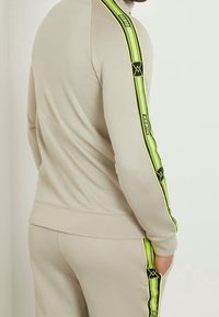 Felpa beige con colletto alto, caratterizzata da audaci strisce nere e verde neon lungo le maniche. Tessuto morbido e design aderente.