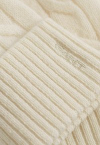 Gebreide trui in roomkleur met geribbelde textuur en een kabelgebreid patroon. Voorzien van subtiele branding in zachte borduursels.