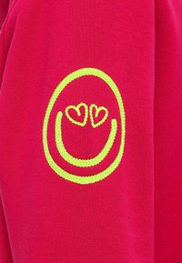 Felpa rossa con un sorriso ricamato di colore giallo neon e occhi a forma di cuore, che presenta una texture morbida e un design informale.