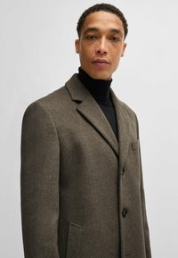 Cappotto di lana verde oliva scuro con rever a punta, tre bottoni e tasche laterali, caratterizzato da un tessuto testurizzato e una vestibilità su misura.