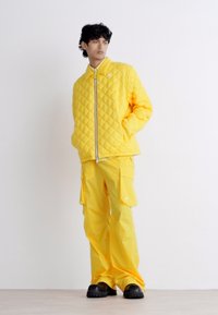Casaco amarelo acolchoado com gola alta, fecho de correr prateado e calças cargo amarelas a condizer. Calçado preto completa o visual.