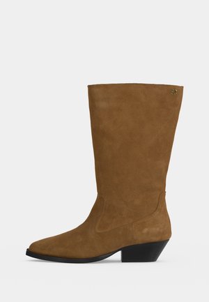 Bota marrón de ante hasta la mitad de la pantorrilla con punta afilada, tacón bajo y inclinado, y pequeño logo dorado cerca del borde superior en el lado exterior.