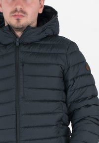 Man in een zwart gewatteerd jack met rits en capuchon, met een klein borstzakje en een ronde logopatch op de bovenarm.