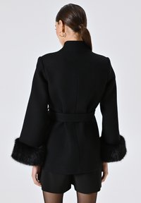 Cappotto nero con tessuto testurizzato, design aderente, maniche lunghe con polsini in pelliccia sintetica e vita con cintura. Visto da dietro.