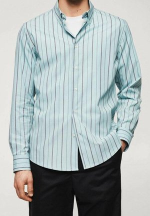 Man die een lichtblauw overhemd met dunne donkere verticale strepen en een zwarte broek draagt, met zijn hand in zijn zak, staand tegen een effen achtergrond.