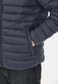 Marintröja i puffer-stil med kviltat mönster, dragkedja fram och sidofickor. Tillverkad av lätt, slätt material med en subtil lyster.