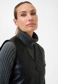 Schwarze Lederweste mit glatter Textur, ausgestattet mit einem tiefen V-Ausschnitt, zwei Knöpfen, getragen über einem grauen, gerippten Rollkragenpullover.
