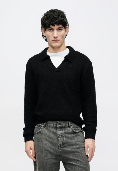 Bărbat tânăr cu păr creț și întunecat, purtând un pulover negru cu guler și mâneci lungi peste o cămașă albă și blugi negri decolorați, stând în picioare pe un fundal simplu.