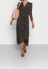 Schwarz gemustertes Midi-Kleid mit langen transparenten Ärmeln, V-Ausschnitt und lockerer Passform. Kombiniert mit einer nude-farbenen Handtasche und schwarzen Riemchen-Heels.
