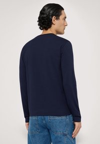 Granatowa koszulka z długim rękawem, wykonana z miękkiej bawełny, z okrągłym dekoltem i czystymi szwami. Model nosi ją z niebieskimi spodniami dżinsowymi.