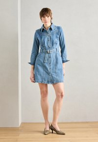 Denim skjortekjole med lange ermer, knappestolpe foran, belted midje og to sidelommer. Lys blå farge med glatt tekstur. Brune høye heler.