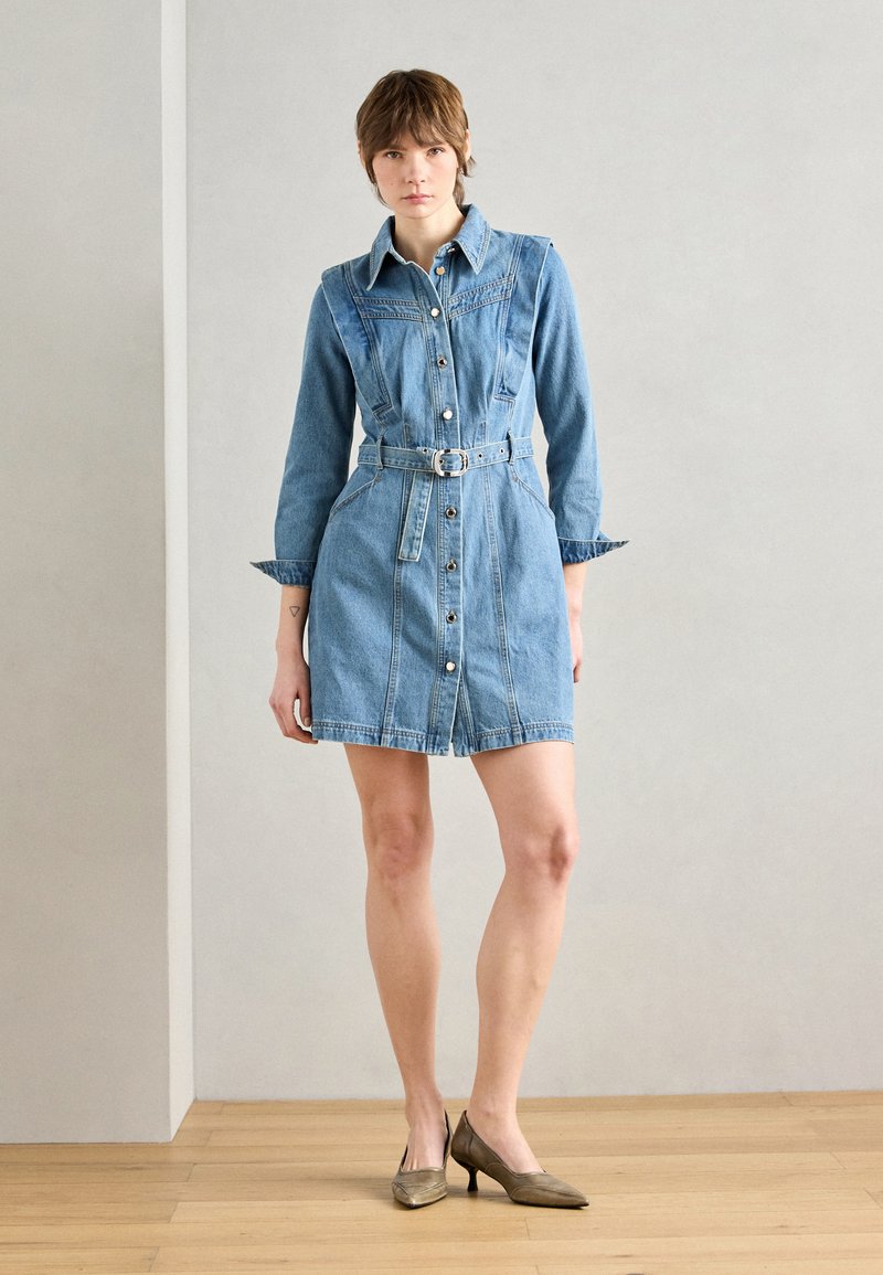 Robe chemise en denim à manches longues, avec un bouton-pression à l'avant, une taille ceinturée et deux poches latérales. Couleur bleu clair avec une texture lisse. Talons bruns.