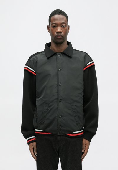 Mustas bomber-jakk läikiva viimistlusega, millel on nööpidega kinnitamine, küljetaskud ja kontrastsete ribadega ribakätised ja alumine äär punase ja valgega.