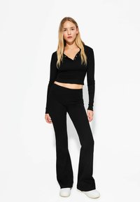 Haut noir à manches longues cropped avec des boutons, associé à un pantalon noir évasé. Tissu lisse avec une coupe ajustée. Baskets blanches complètent le look.