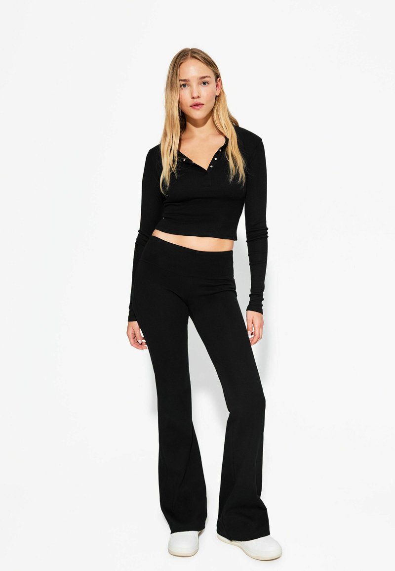 Haut noir à manches longues cropped avec des boutons, associé à un pantalon noir évasé. Tissu lisse avec une coupe ajustée. Baskets blanches complètent le look.