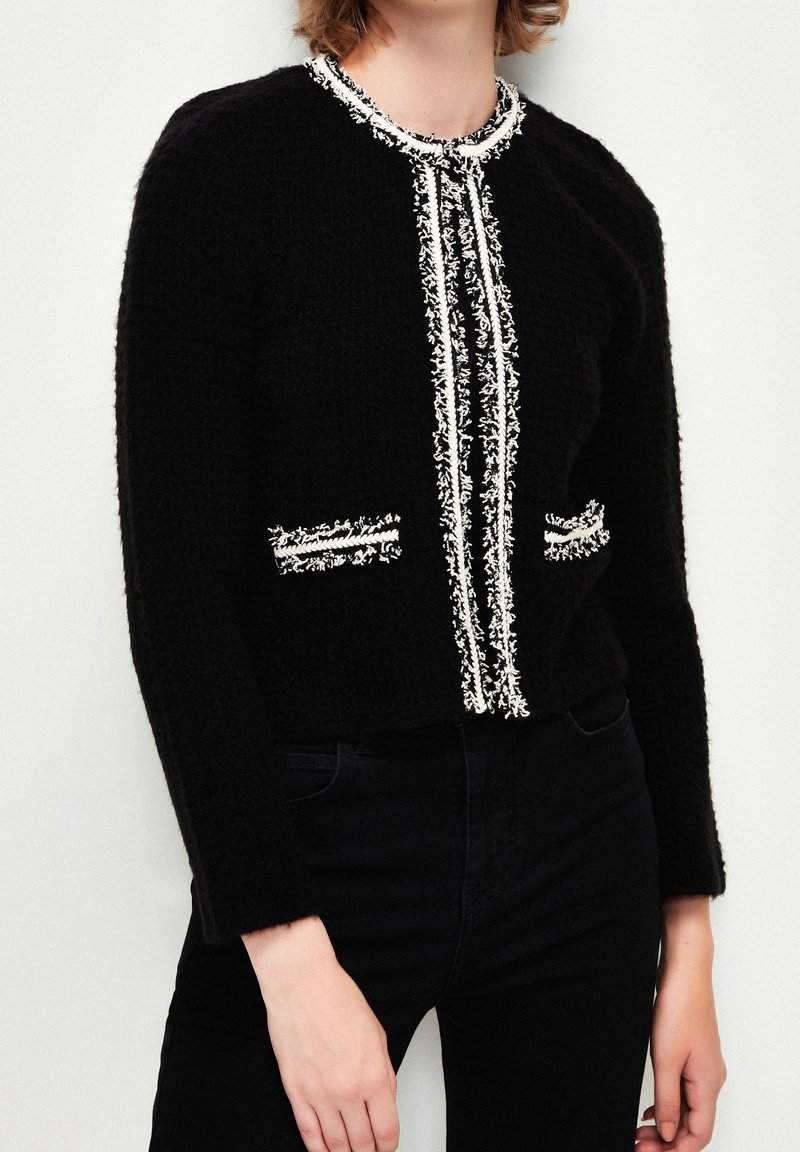 Personne portant un cardigan noir texturé avec une bordure à franges blanches le long du devant, de l'encolure et de deux petites poches avant, associé à un pantalon noir.