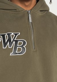 Sudadera color verde oliva con diseño de media cremallera, presenta las letras bordadas en blanco y negro "WB" en el pecho y una tela suave y texturizada.