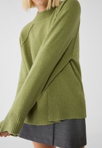 Grüner Strickpullover mit strukturiertem Finish, geripptem Halsausschnitt und Saum sowie überschnittenen Schultern. Kombiniert mit einem grauen Rock.