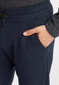 Marina blå joggingsbyxor gjorda av texturerad fleece, med en dragsko i midjan och sidofickor, där en hand vilar i en ficka.