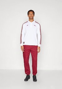 Balts garu piedurkņu polo ar sarkaniem svītrām un logotipu, kopā ar sarkaniem biksēm. Melni apavi ar teksturētām zolēm. Vienkāršs, sportisks dizains.