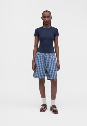 Kvinde står, iført en navy tætsiddende kortærmet skjorte, blå-hvide ternede knælange shorts, hvide sokker og brune sneakers.