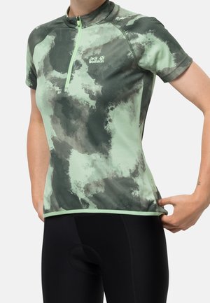 Kvinde justerer lysegrøn og mørkegrå camouflage-cykeltrøje med korte ærmer, frontlynlås og Jack Wolfskin-logo, iført sorte cykelshorts.