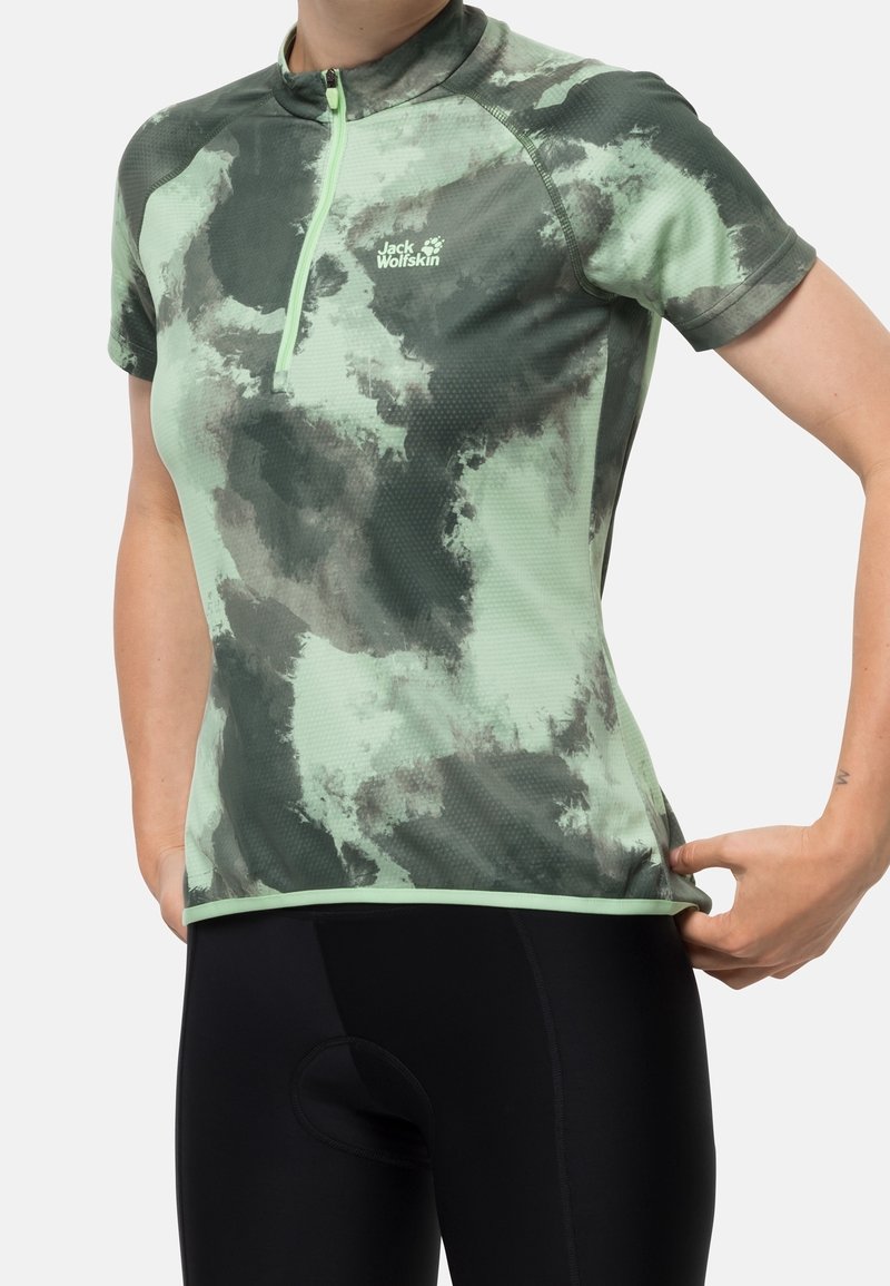 Mujer ajustándose un maillot de ciclismo de manga corta con estampado de camuflaje en verde claro y gris oscuro, con cremallera frontal y logo de Jack Wolfskin, llevando pantalones cortos de ciclismo negros.