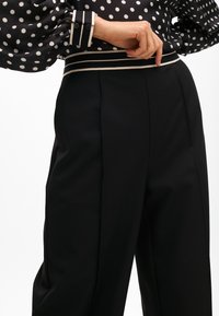 Black trousers with a wide-leg cut, featuring pleats and a textured waistband with contrasting stripes. Smooth fabric, tailored fit.