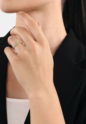 Goldfarbiger Ring mit klaren Edelsteinen im Wellenmuster. Hand in der Nähe des Halses positioniert, vor einem schwarzen Blazer-Hintergrund.