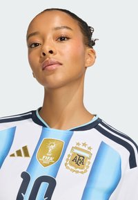 Mladá žena v dresu argentinského fotbalu s odznaky Mistrovství světa FIFA 2022 a AFA, sebevědomě hledící do kamery.