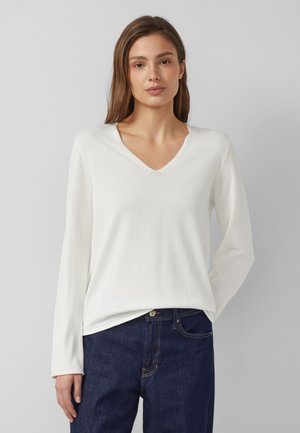 Frau mit glatten braunen Haaren, die ein weißes, langärmliges V-Ausschnitt-Oberteil und dunkelblaue Jeans trägt und vor einem schlichten grauen Hintergrund steht.