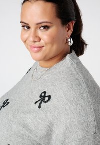 Femme aux yeux marron et cheveux foncés tirés en arrière, portant des boucles d'oreilles anneaux argentées, des colliers argentés superposés et un pull gris avec des motifs de rubans noirs.