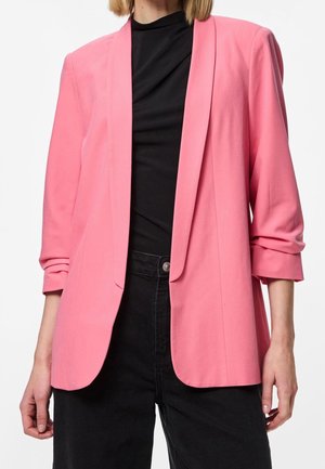 Person trägt ein leuchtend pinkes Sakko über einem schwarzen Oberteil mit schwarzer, hochtaillierter Hose, steht vor einem weißen Hintergrund.