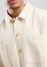 Lichtbeige overhemden gemaakt van dikke katoen, met twee zakken aan de voorkant en gele knopen. Close-up van de kraag en de knopenlijst.