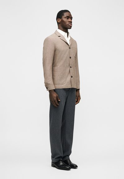 Άνδρας που φοράει μπεζ σακάκι με κουμπιά, λευκό πουκάμισο, γκρι παντελόνι και μαύρα επίσημα παπούτσια, στέκεται μπροστά σε ένα απλό ανοιχτόχρωμο φόντο.